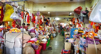 A busy covered market aisle, vendors seated behind baskets of produce, dried fish and garlands of marigold